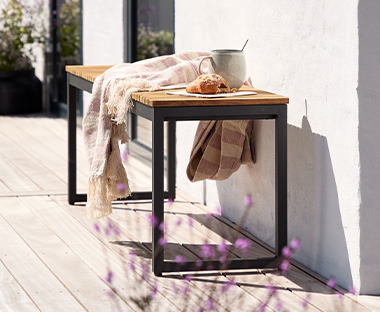 Moderna baštenska klupa na dvorištu uz bijeli zid i deka, kroasan i šolja čaja na njoj