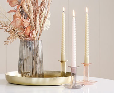 Tri spilarne svijeće na malim staklenim svijećnjacima pored vaze sa cvijećem na poslužavniku