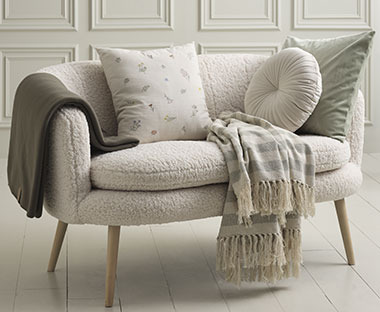 Bijela čupava sofa sa ukrasnim jastucima u bež tonovima i dekom sa bež i sivim crtama