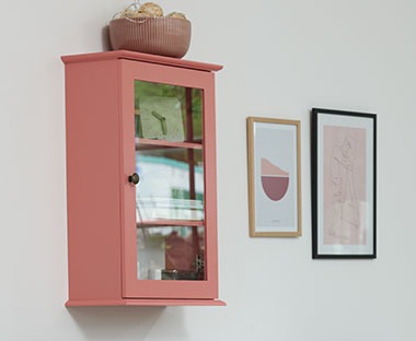 Ormarić u roze boji montiran na bijeli zid pored dva okvira za slike