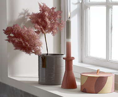 Svijeća, ukrasna kutija, svijećnjak i umjetna biljka u roze nijansama na prozoru