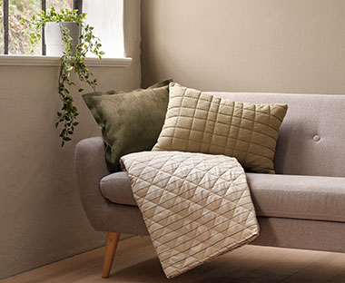 Kauč sa ukrasnim jastucima i štepanim dekama pored prozora sa umjetnom biljkom u sivoj saksiji
