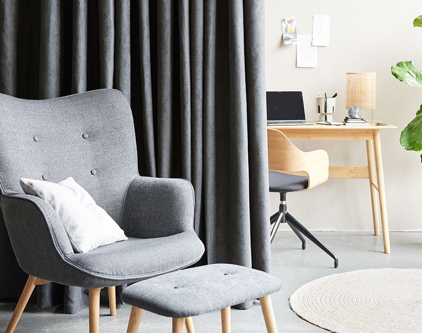 Sive zavjese razdvajaju home office sa stolom i radnom stolicom od dnevne sobe sa foteljom