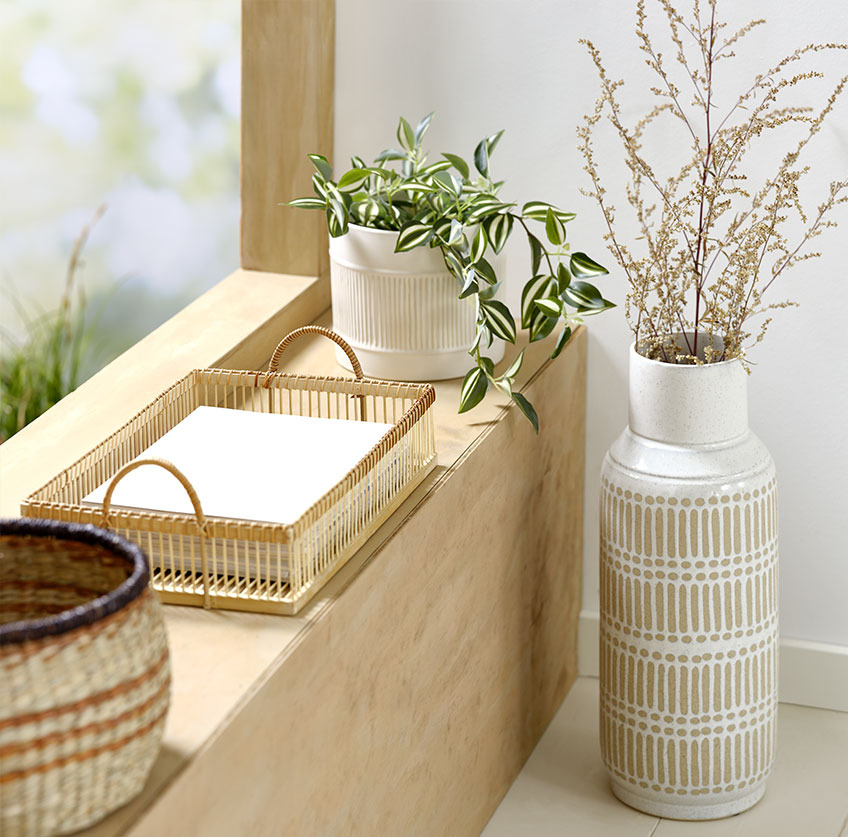 Visoka vaza pored prozorske daske sa poslužavnikom od bambusa i bijelom saksijom sa umjetnom biljkom