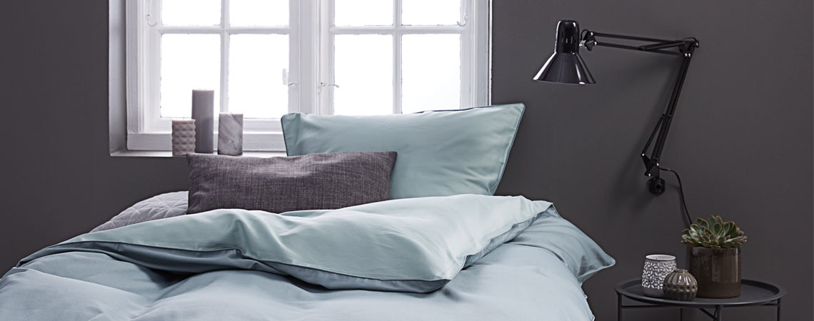 Krevet i svijetlo plava posteljina ispod prozora i lampa na noćnom ormariću