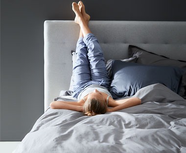 Žena se odmara na krevetu sa rukama ispod glave i nogama podignutom na uzglavlje