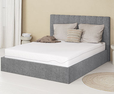 Sivi tapacirani okvir bračnog kreveta i kvalitetan bijeli madrac od pjene sa jastucima na njemu