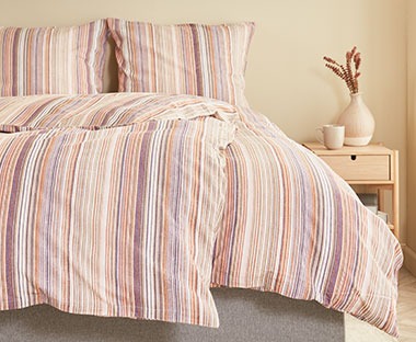 Roze posteljina na pruge na krevetu i elegantni noćni ormarić od svijetlog drveta pored