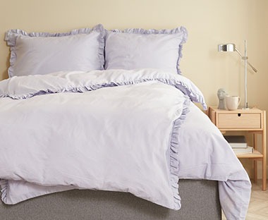 Svjetlo ljubičasta posteljina na krevetu pored noćnog ormarića od svijetlog drva